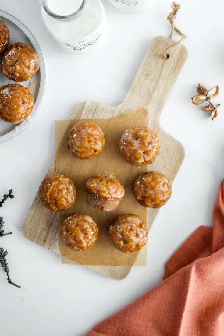 Overhead view of glazed pumpkin donut holes on a wooden kitchen board, jug of milk, additional donuts holes on a tray on a white countertop.