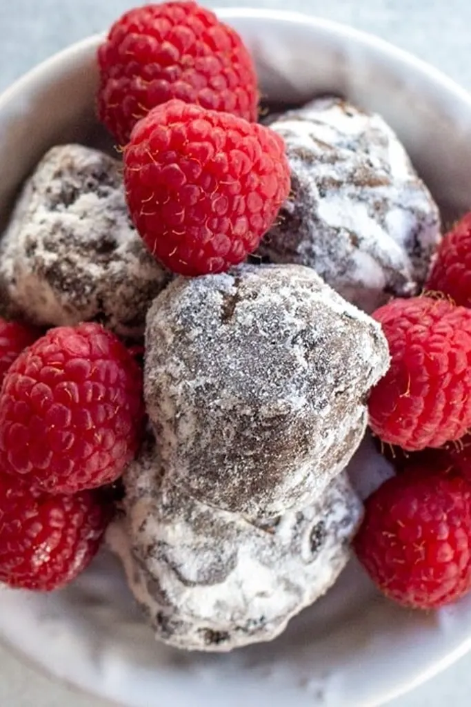 truffle shaped dark chocolate cookies with powder and raspberries on top in white bowl 