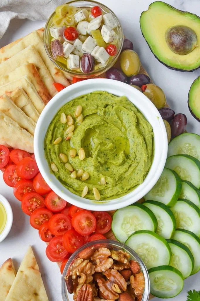avocado hummus in white bowl with pita, tomatoes and cucumbers on side 