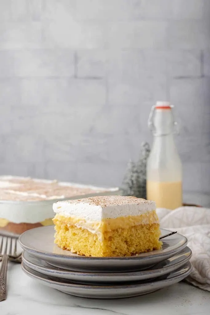 slice of eggnog poke cake with icing on stack of grey plates 