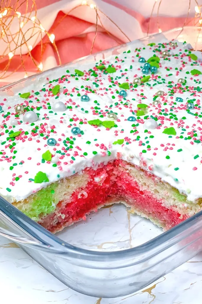 Christmas colored poke cake with white icing and Christmas sprinkles on top 