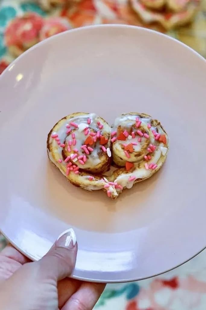 25 Valentine’s Day Breakfast Ideas For Kids 6 iced heart shaped cinnamon rolls with pink and heart sprinkles on white plate