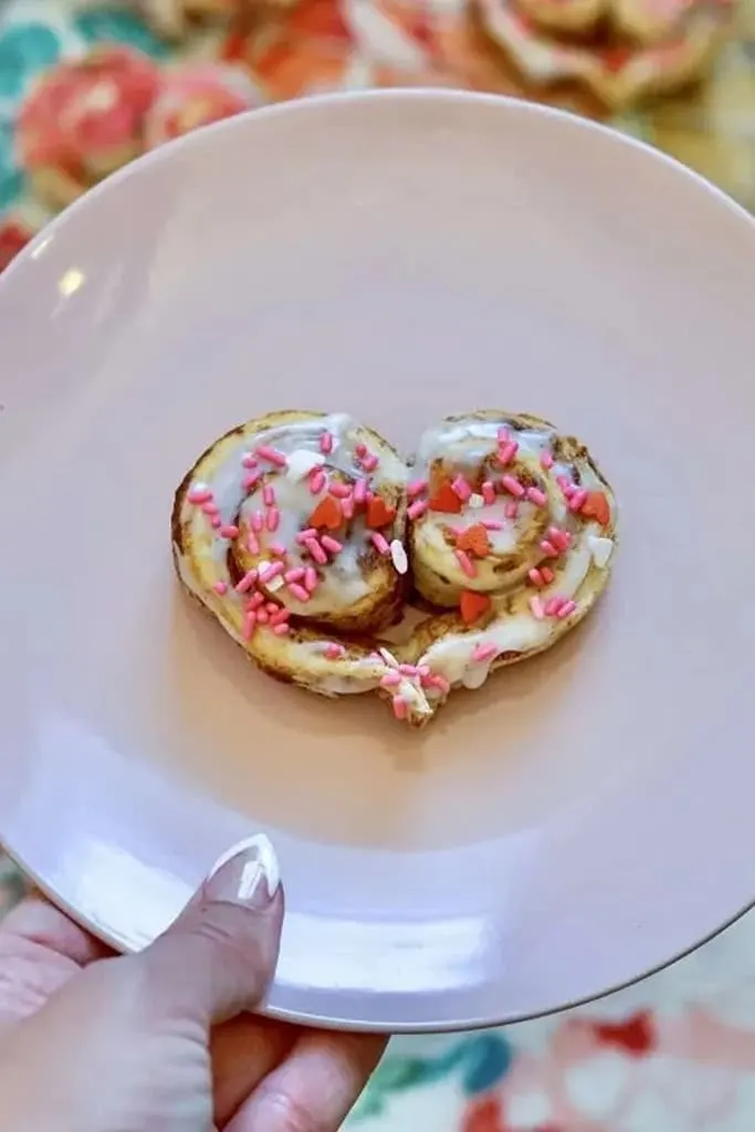 iced heart shaped cinnamon rolls with pink and heart sprinkles on white plate 