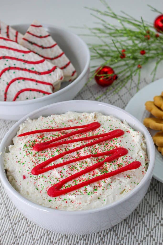 Little Debbie Christmas Tree Cake Dip (Easy No Bake Christmas Dessert) 4 little debbie christmas tree cake dip in bowl with red frosting drizzle in background is christmas greenery and little debbie christmas tree cakes
