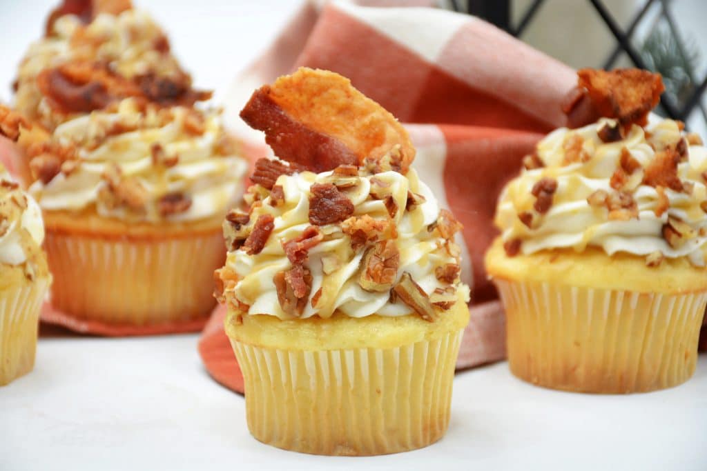 Maple Bacon Cupcakes with Whiskey Caramel Sauce 6 close up of Maple Bacon Bourbon Cupcakes in white cupcake liners with bacon on top with more in background