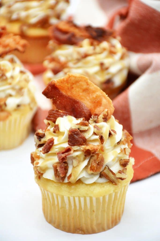 close up of maple bourbon cupcake in white cupcake cup with bacon on top
