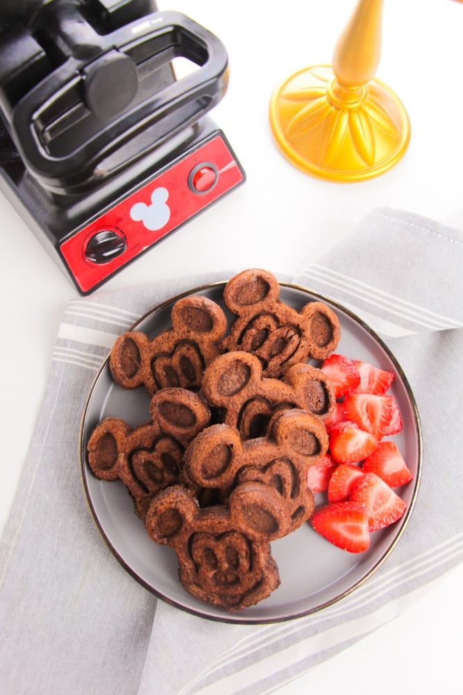 Chocolate Mickey Mouse Waffles 11 overhead view white plate of cooked Mickey Mouse waffles and strawberries on the side and waffle maker in background