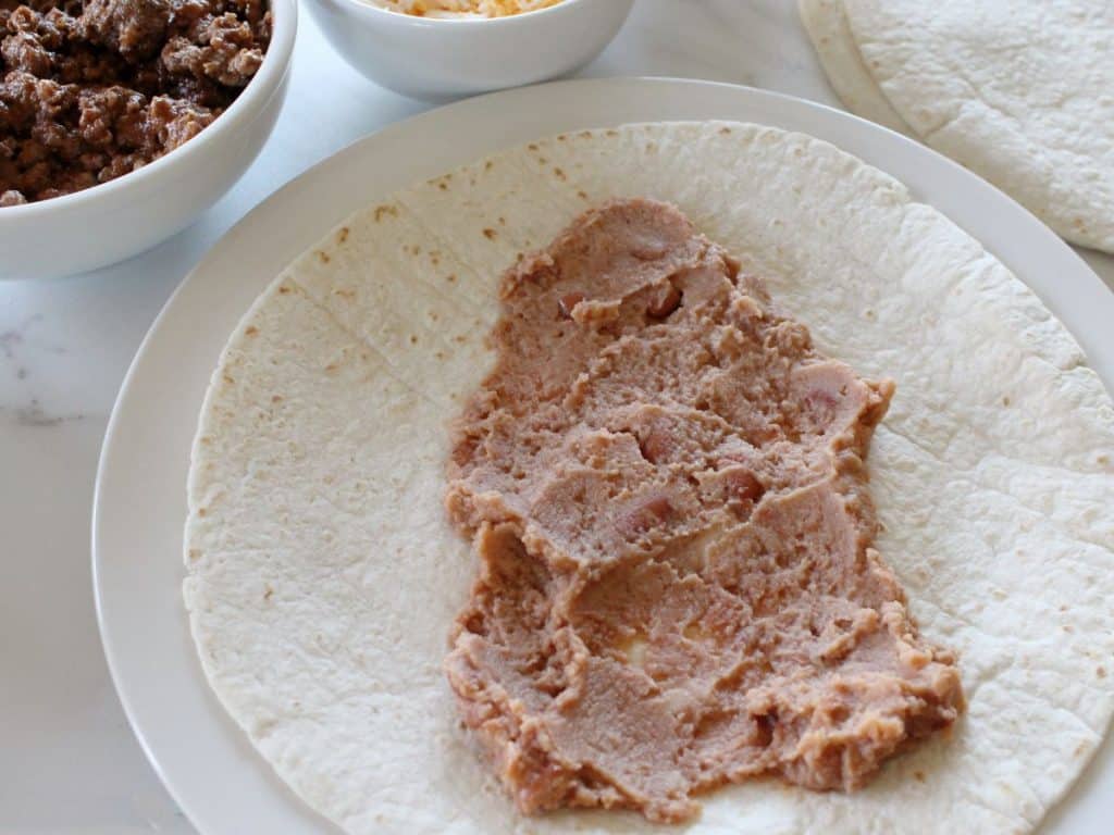 Taco Bell Enchrito Copycat Recipe 6 refried beans spread on a tortilla on a white plate