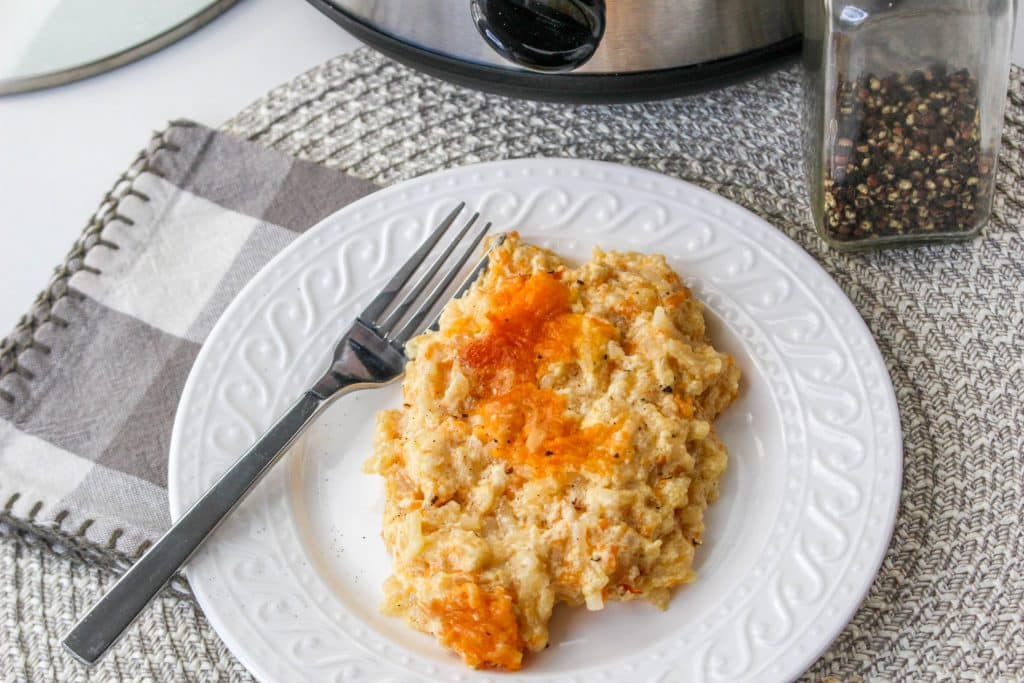 overhead view of slow cooker cheesy hash brown casserole in slow cooker on white plate with silver spoon and crockpot in background