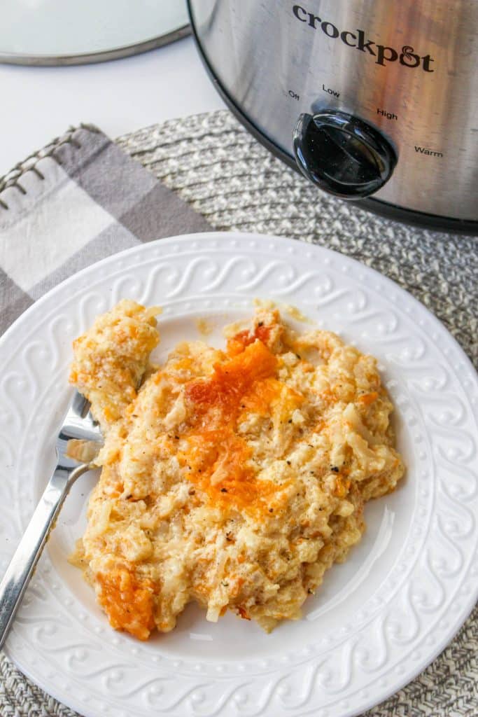 Slow Cooker Cracker Barrel Cheesy Hash Brown Casserole 4 overhead view of cooked hash brown casserole in slow cooker on white plate with silver spoon and crockpot in background