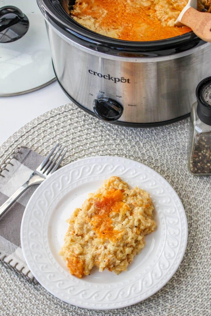 Slow Cooker Cracker Barrel Cheesy Hash Brown Casserole 3 overhead view of cooked hash brown casserole in slow cooker on white plate with silver spoon and crockpot in background