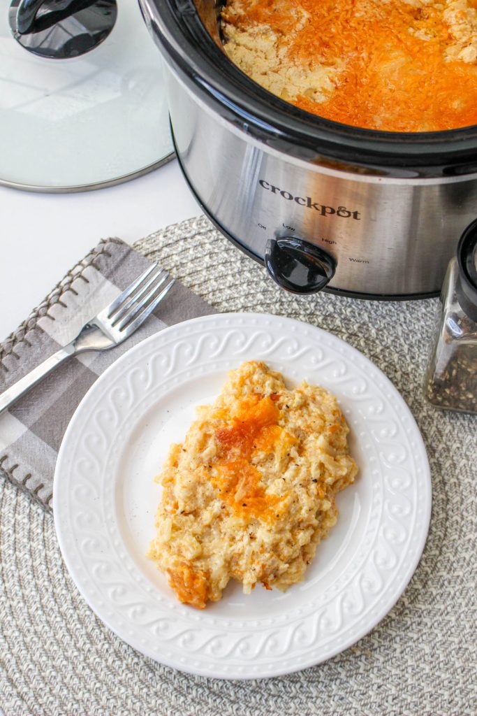 Slow Cooker Cracker Barrel Cheesy Hash Brown Casserole 8 overhead view of cooked hash brown casserole in slow cooker on white plate with silver spoon and crockpot in background