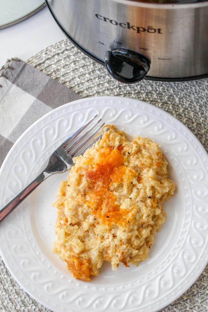 overhead view of cooked hash brown casserole in slow cooker on white plate with silver spoon