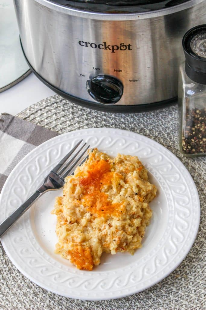Slow Cooker Cracker Barrel Cheesy Hash Brown Casserole 6 overhead view of cooked hash brown casserole in slow cooker on white plate with silver spoon and crockpot in background