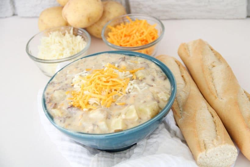 Sausage Potato Soup in Crockpot 3 Sausage potato soup in a blue bowl with glass bowls holding shredded cheese for toppings. Long crusty bread pieces are laying on the white counter beside the bowl.