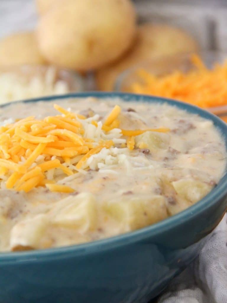 Sausage Potato Soup in Crockpot 4 Close up of creamy sausage potato soup with shredded cheese on top. The soup is in a blue bowl and there are potatoes in the background sitting on a white surface.
