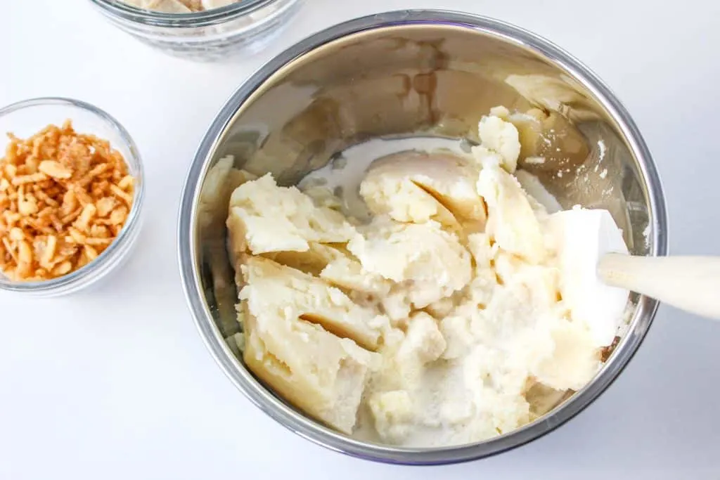 Mixing bowl with leftover mashed potato and half and half with mixing spoon on a white countertop.