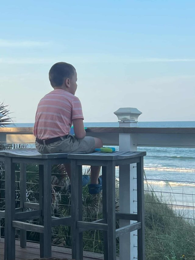 A young boy sits on a high wooden table overlooking the ocean, gazing at the waves under a clear sky.