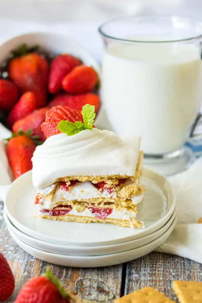 A slice of strawberry icebox cake on stacked white plates, glass of creamy milk and bowl of fresh strawberries in the background.