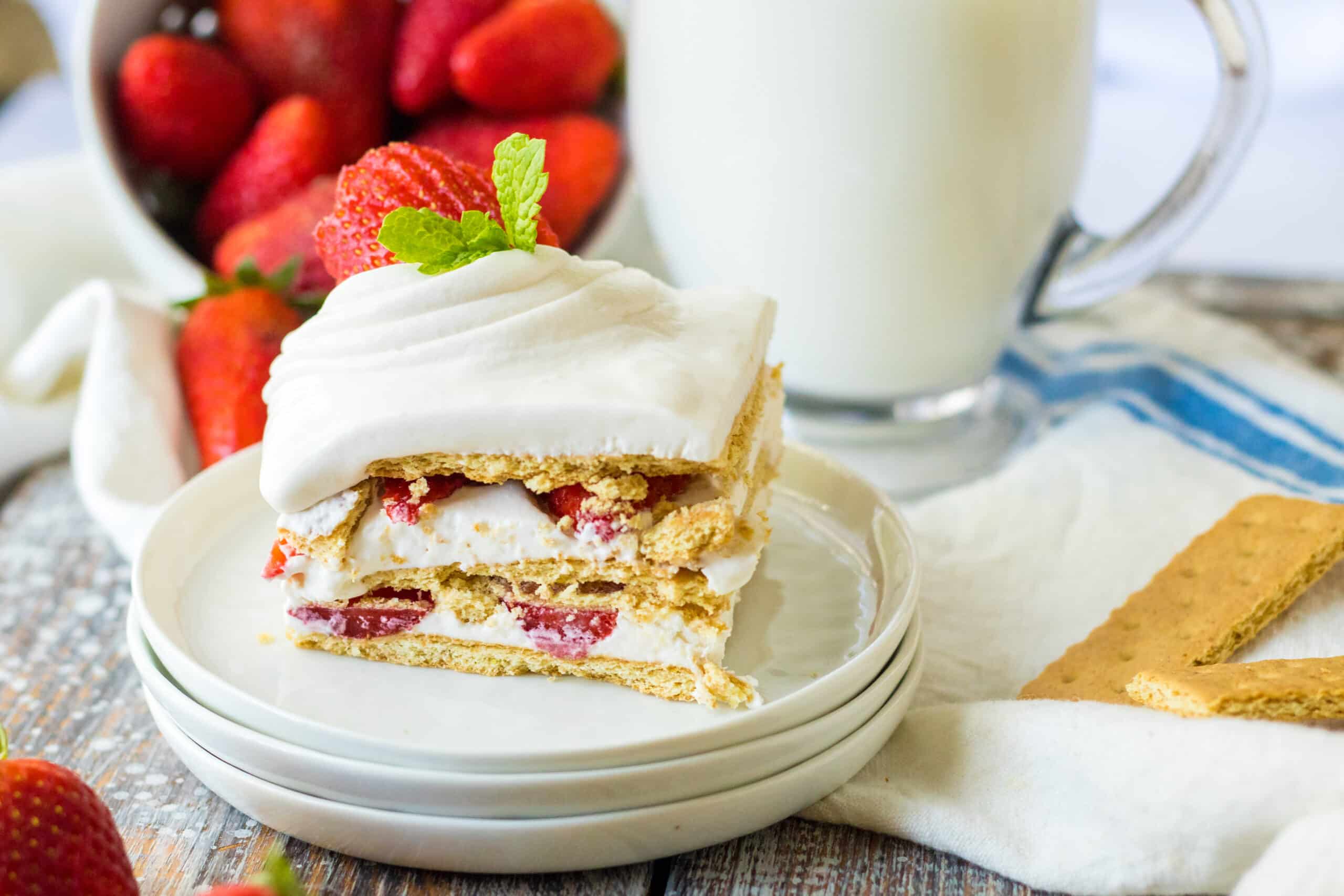 Strawberry ice box cake on white place with strawberries in background, glass of milk and loose Graham crackers.