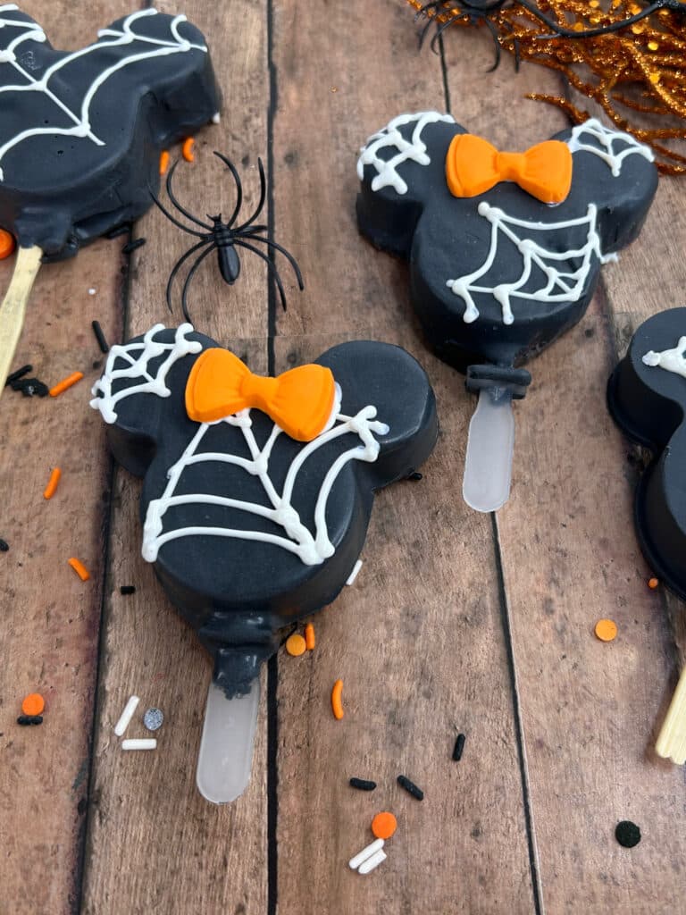 Disney Halloween Recipe (Mickey Oreo Pops) 3 overhead view of black halloween mickey Oreo pops with white webbing and orange Minnie bow