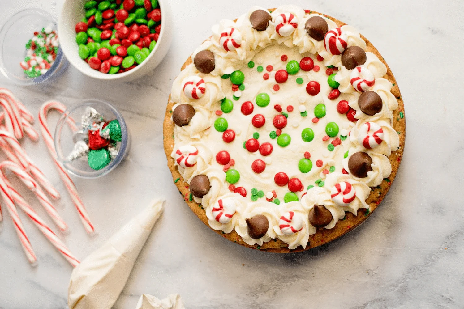 Overhead view of Christmas cookie cake topped with vanilla frosting and candy, vanilla frosting in a pipping bag and small bowls of candy on a marble countertop.