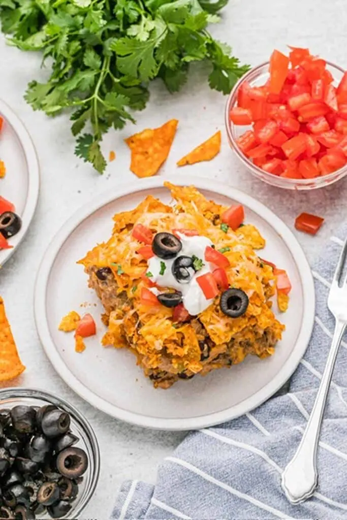 Serving of Dorito casserole on a white plate, bowl of black olives, bowl of diced tomatoes.