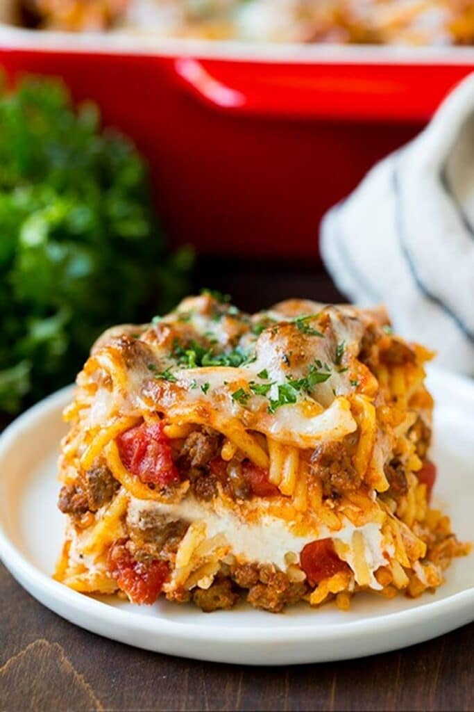 25 Venison Burger Casserole Recipes 7 Serving of Million dollar spaghetti on a white plate, remainder of casserole in a red casserole dish in the background.