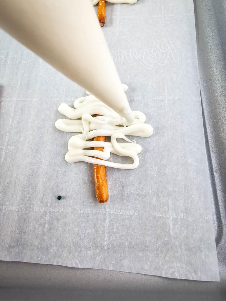 Pretzel Christmas Trees 7 A piping bag drizzling melted white chocolate into a pretzel stick on a parchment paper lined tray.