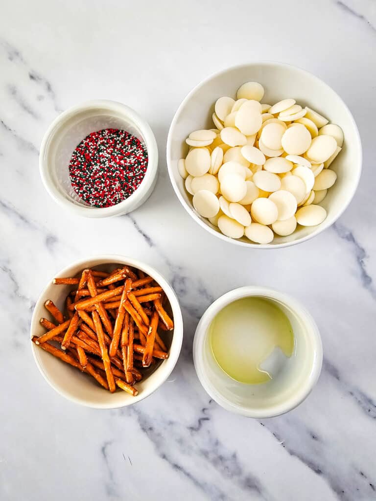 Pretzel Christmas Trees 3 Flat lay shot of ingredients for the Pretzel Christmas Trees. These include melting white chocolate wafers, pretzel sticks, olive oil and nonpareils, all in separate white bowls. The ingredients are then placed on a marble countertop.