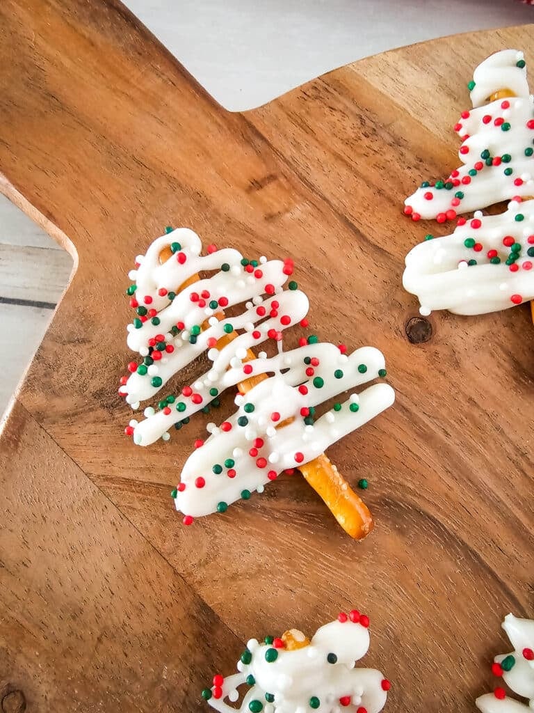 Pretzel Christmas Trees 2 Closeup shot of a Pretzel Christmas Tree on a round wooden board. The holiday treat is made of a pretzel stick, then drizzled with melted white chocolate to mimic the shape of a pine tree. Then it's sprinkled with red, green and white sprinkles. There are several other Pretzel Christmas Trees surrounding the treat.