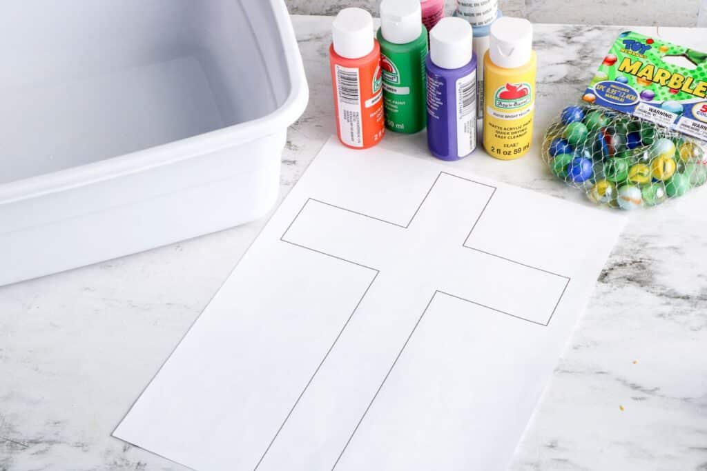 Cross Marble Painting Craft for Kids 16 Materials needed to make the Cross Marble Painting such as the rectangular plastic bin, assorted acrylic paints in plastic bottles, a small bag of marbles, and the printed cross template, all placed on a marble surface.