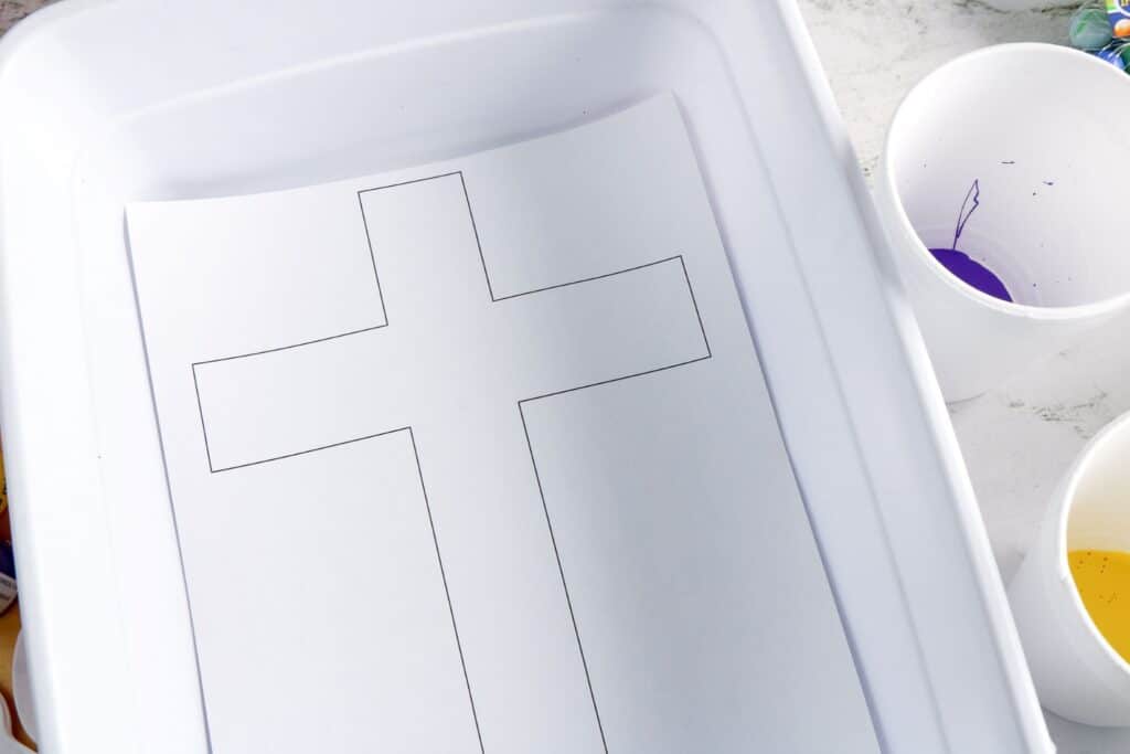 Cross Marble Painting Craft for Kids 17 A sheet of white paper with the printed cross template in it. The printed template is then placed on a rectangular plastic box with paints in cups beside it.