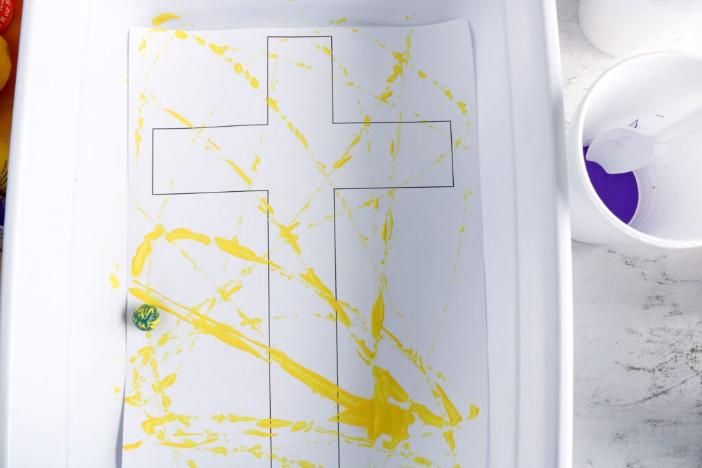 Cross Marble Painting Craft for Kids 20 The blue marble coated in yellow paint, leaving streaks of yellow paint across the printed cross template.