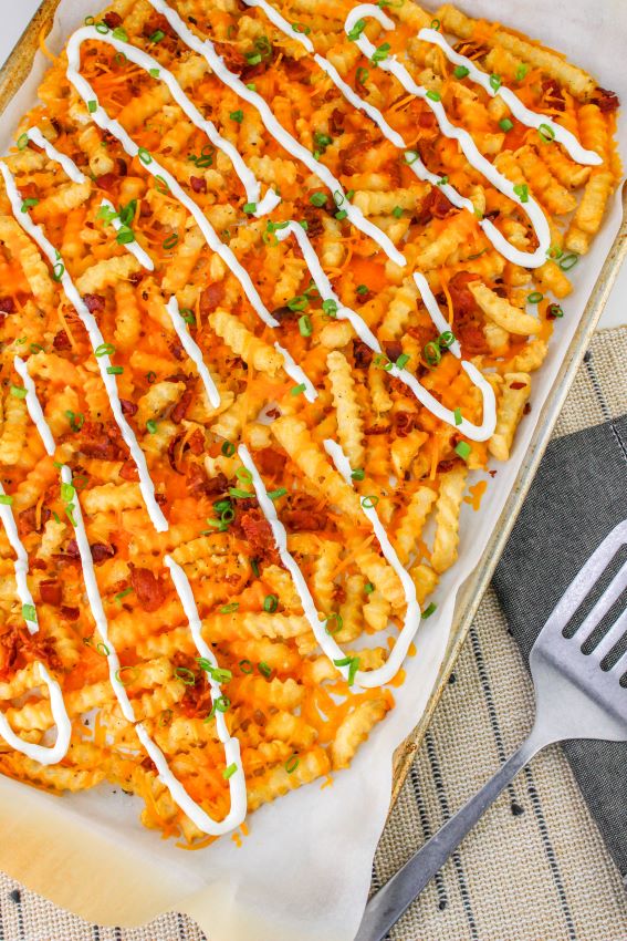 Loaded French Fries With Bacon 21 Flat lay, partial shot of the finished Loaded French Fries With Bacon on a sheet pan lined with parchment paper. Beside it is a stainless steel spatula on a table with burlap cloth.