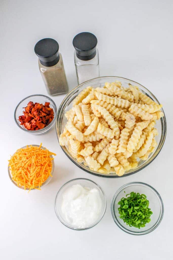 Loaded French Fries With Bacon 23 Flat lay shot of the ingredients needed to make the Loaded French Fries With Bacon. Ingredients include frozen fries in a large glass bowl, cooked bacon bits, shredded cheese, sour cream, chopped green onions, salt and pepper, all on a white surface.