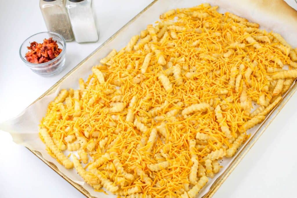Loaded French Fries With Bacon 25 High-angle shot of a sheet tray lined with parchment paper, with baked fries topped with shredded cheese.