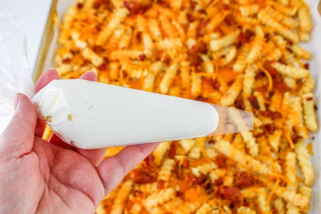 Loaded French Fries With Bacon 27 Focus shot on a hand holding a plastic piping bag with sour cream over a tray of the Loaded French Fries With Bacon on a parchment paper.