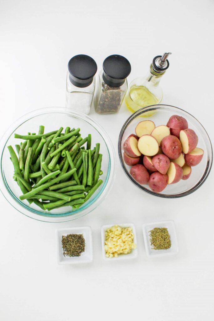 Leftover Roasted Potatoes and Green Beans 4 Overhead shot of the ingredients for the Leftover Roasted Potatoes and Green Beans such as chopped green beans, halves potatoes, garlic, salt, pepper and herbs, in separate containers, all placed on a white surface.