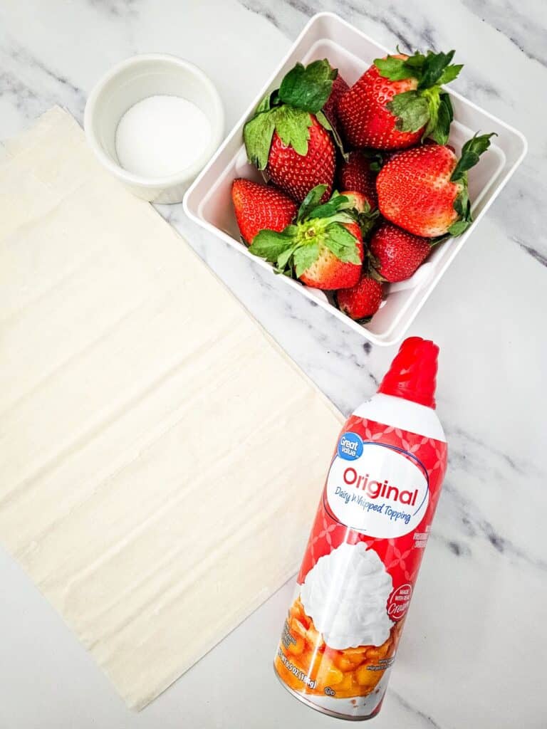 Flat lay shot of ingredients for the Mini Strawberry Shortcake Bites such as phyllo pastry, fresh strawberries, white sugar on a white ramekin and a can of whipped cream.