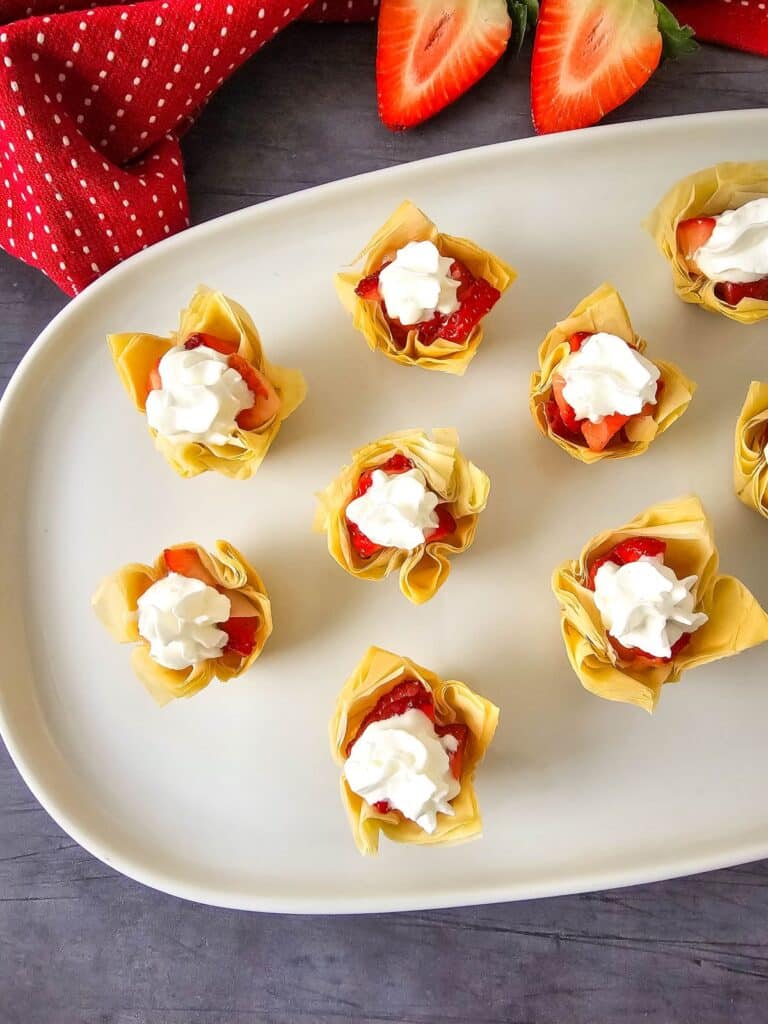 Flat lay shot of the finished batch of Mini Strawberry Shortcake Bites on a white rectangular platter with rounded corners. Beside the platter are a red polka dot table napkin and fresh strawberries cut in half.