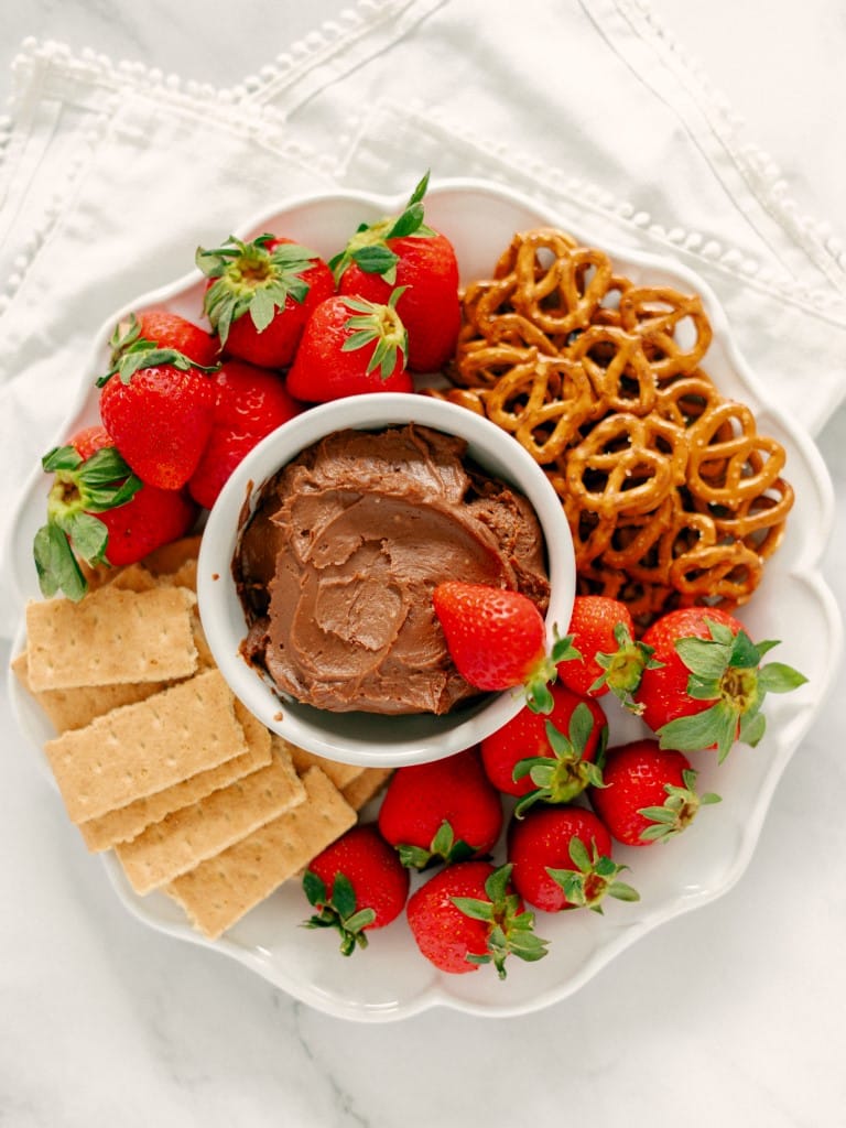 Flat shot of the finished Nutella Fruit Dip in a white bowl, placed in the middle of a platter and surrounded with assorted dippables such as fresh strawberries, graham crackers, and baked mini pretzels.