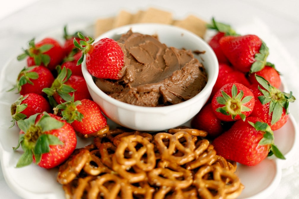 A decadent serving of the Nutella Fruit Dip on a white bowl, topped with a single fresh strawberry. The white bowl is then placed on a platter surrounded by assorted dippables such as pretzels, fresh strawberries and wafer squares.