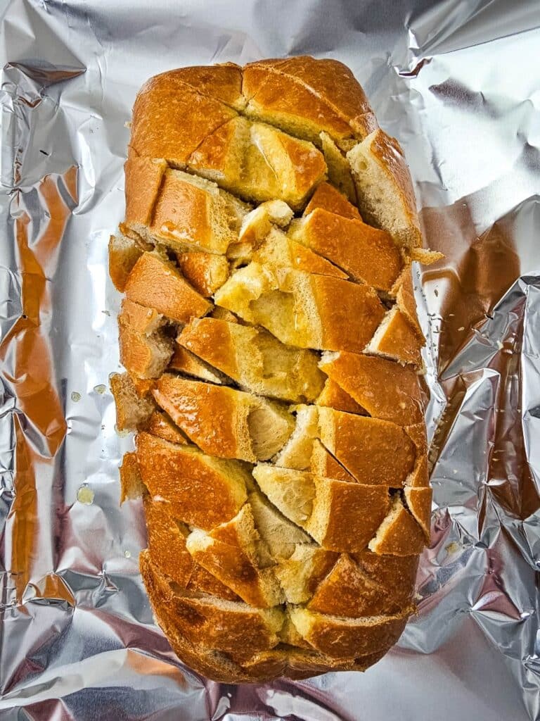 Flat lay shot of a loaf or sliced bread, with criss-cross slices on its surface and then brushed with butter. The loaf is on a prepared sheet tray.