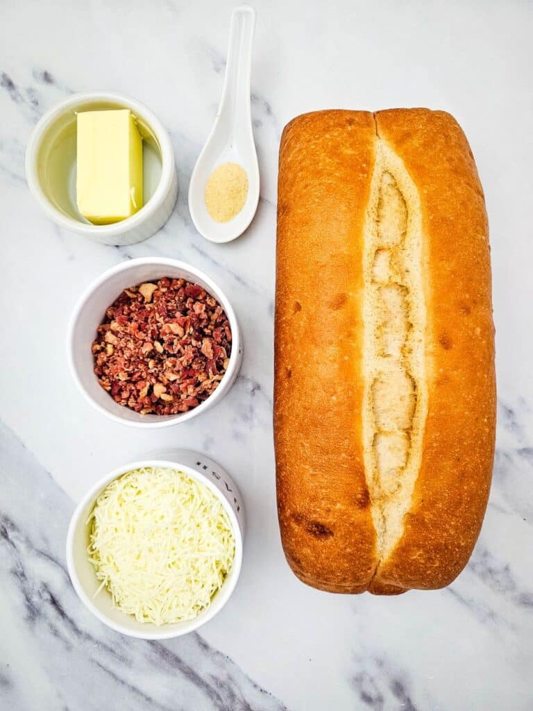 Flat lay shot of ingredients such as a loaf of bread, bacon bits, shredded cheese, butter and garlic powder. The toppings ingredients are on separate white bowls while the loaf is on a marble surface.