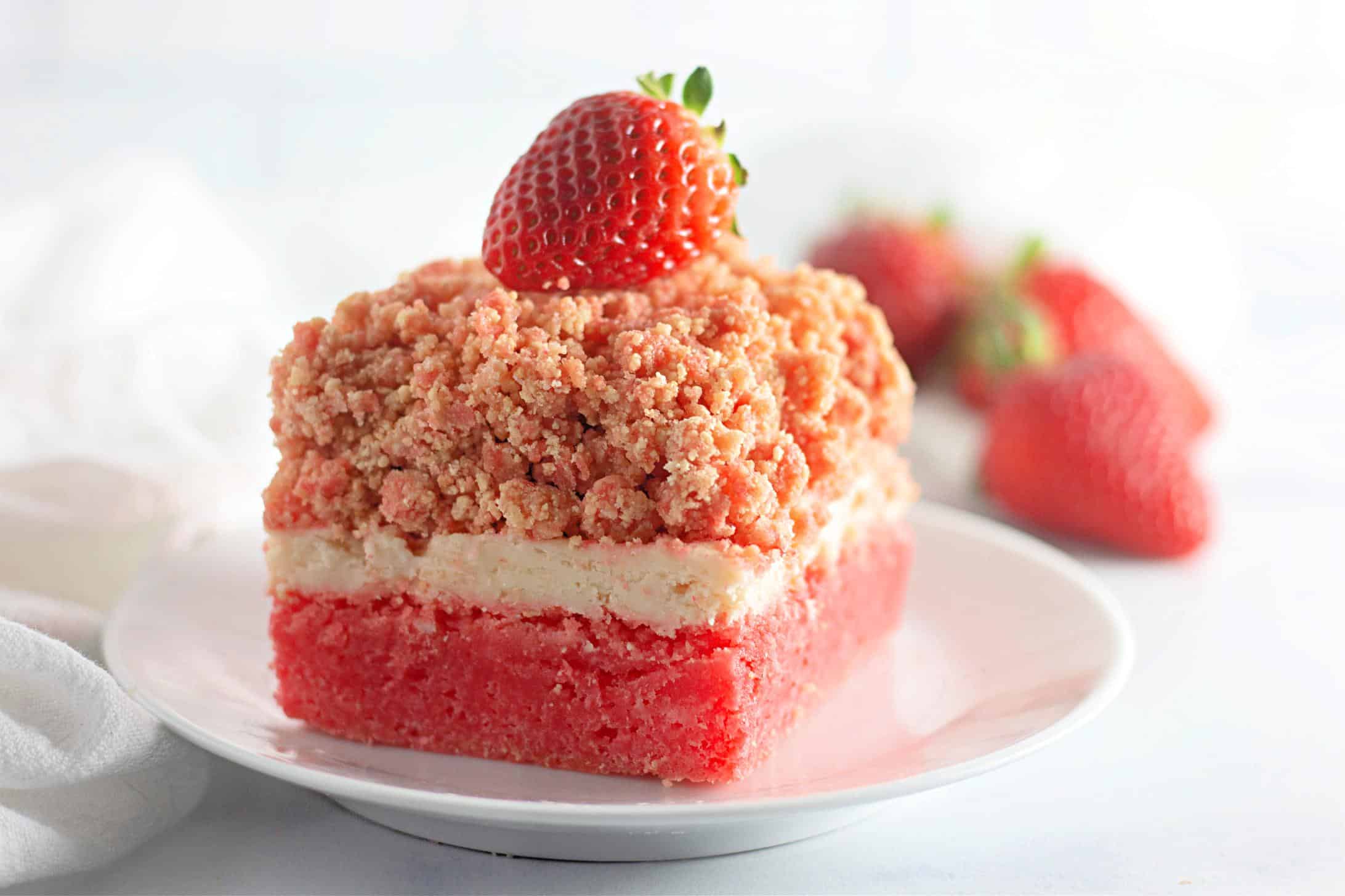 Focus shot on a square slice of the finished Easy Strawberry Crunch Cake Bar. The dessert is topped with a single fresh whole strawberry, then placed on a white plate.