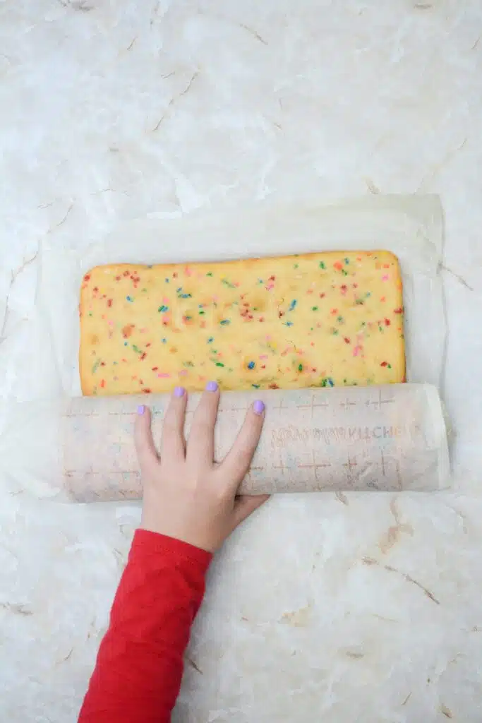 Easy From Scratch Confetti Birthday Cake Roll Recipe with Cream Cheese Frosting 21 A manicured hand rolling up the sheet cake with the parchment paper over each rolled layer of the cake.