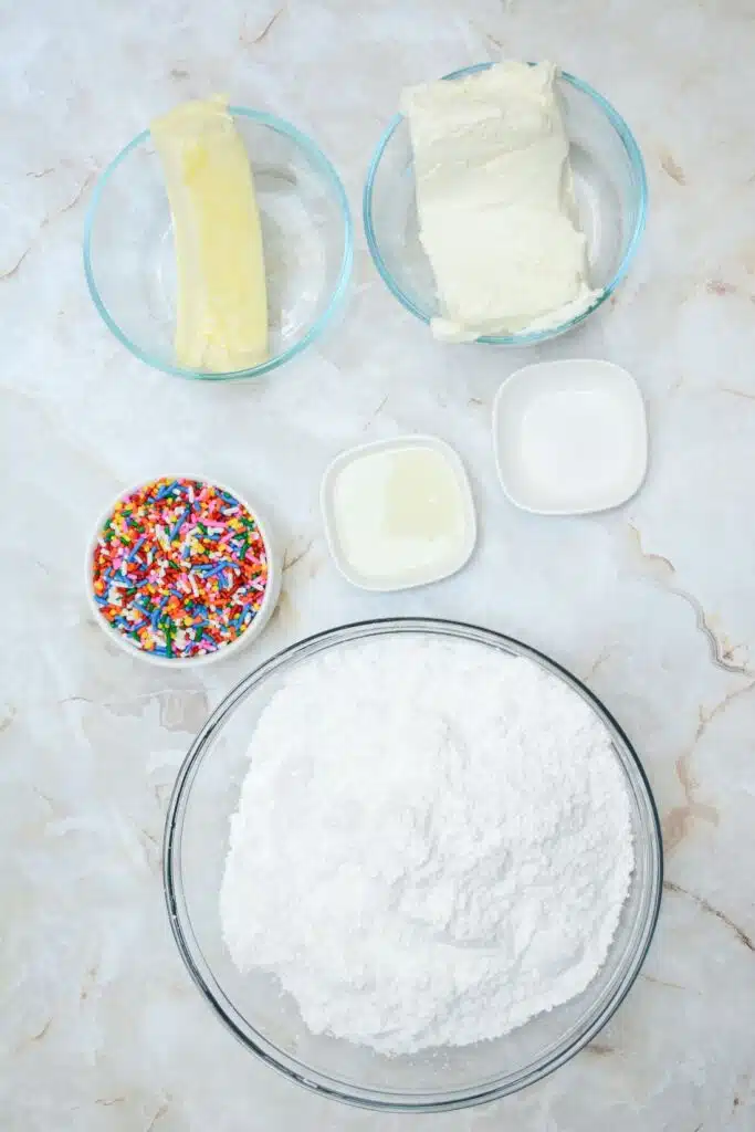 Easy From Scratch Confetti Birthday Cake Roll Recipe with Cream Cheese Frosting 10 Flat lay shot of the ingredients for the cream cheese frosting in separate glass bowls, all placed on a marble surface.