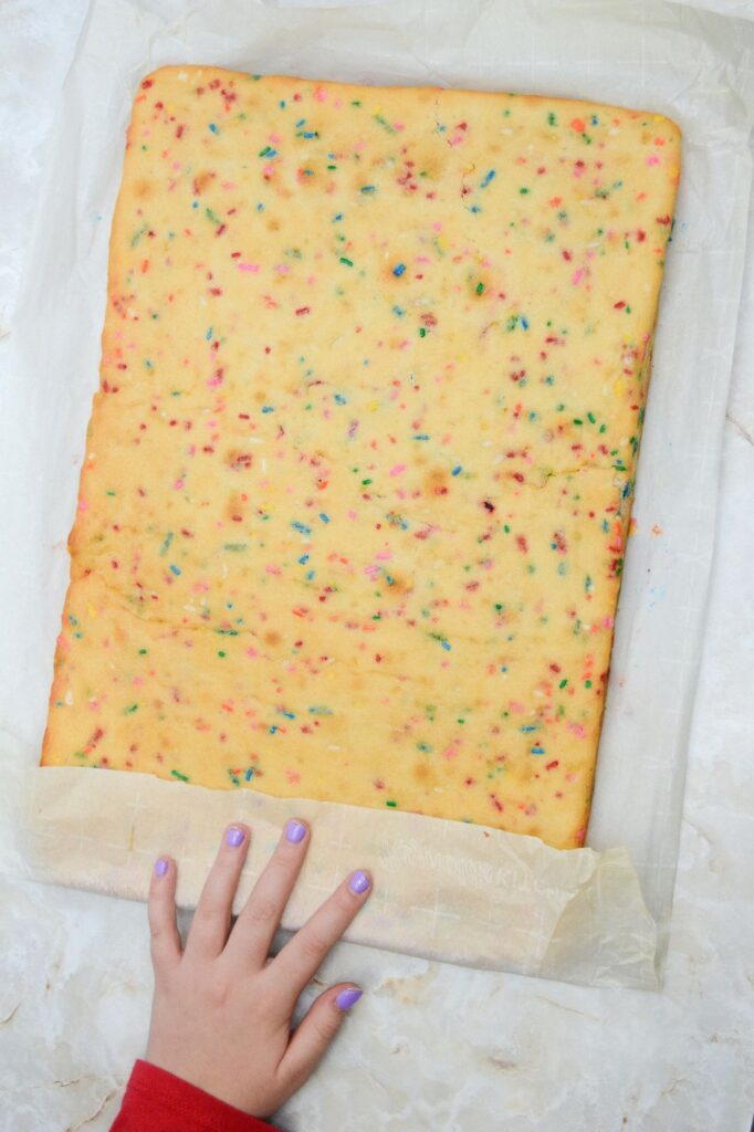 A manicured hand folding the lower flap or the parchment paper over the lower section of the baked sheet cake placed on top of it.