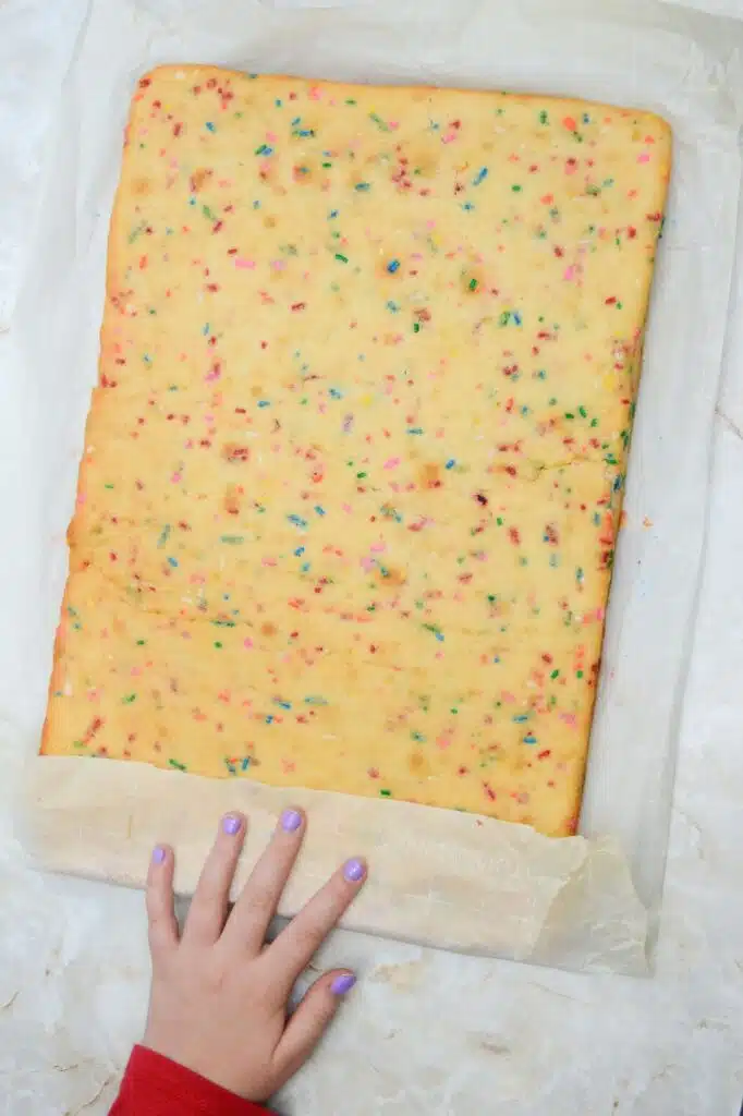 Easy From Scratch Confetti Birthday Cake Roll Recipe with Cream Cheese Frosting 20 A manicured hand folding the lower flap or the parchment paper over the lower section of the baked sheet cake placed on top of it.